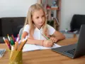 Dziewczynka z blond włosami pisze w zeszycie przy laptopie, obok stoją kolorowe kredki.