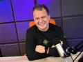 Uśmiechnięty mężczyzna w czarnej bluzie siedzi przy biurku z mikrofonem, na tle fioletowej ściany.