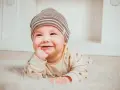 Uśmiechnięte dziecko w czapce leży na białym futerku, wkładając palec do buzi.