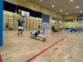 Dzieci ćwiczą siatkówkę na sali gimnastycznej, wykonując pompki i podbijając piłkę.