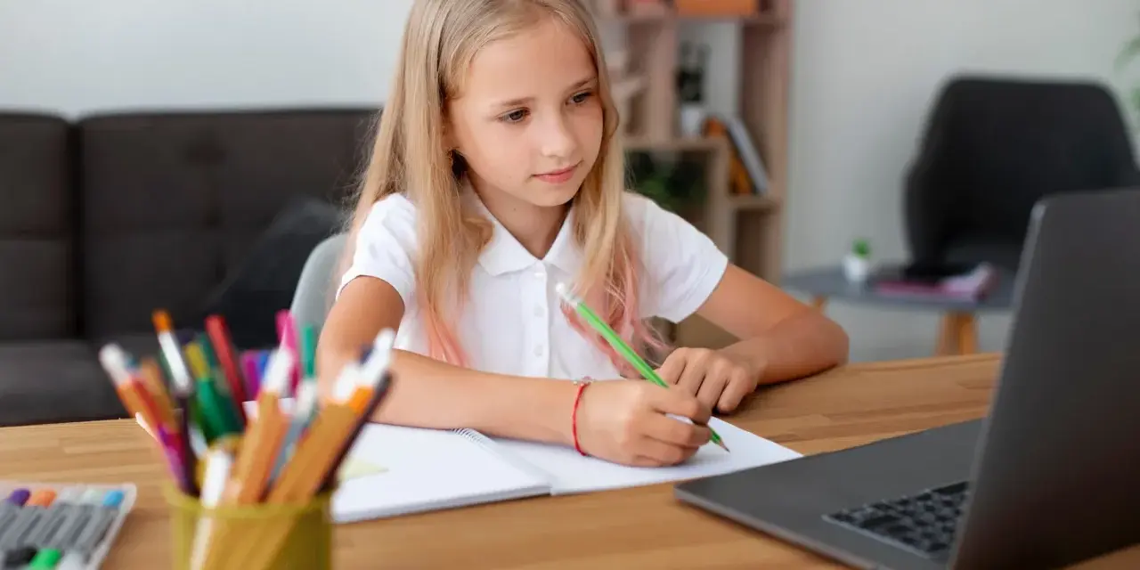 Dziewczynka z blond włosami pisze w zeszycie przy laptopie, obok stoją kolorowe kredki.