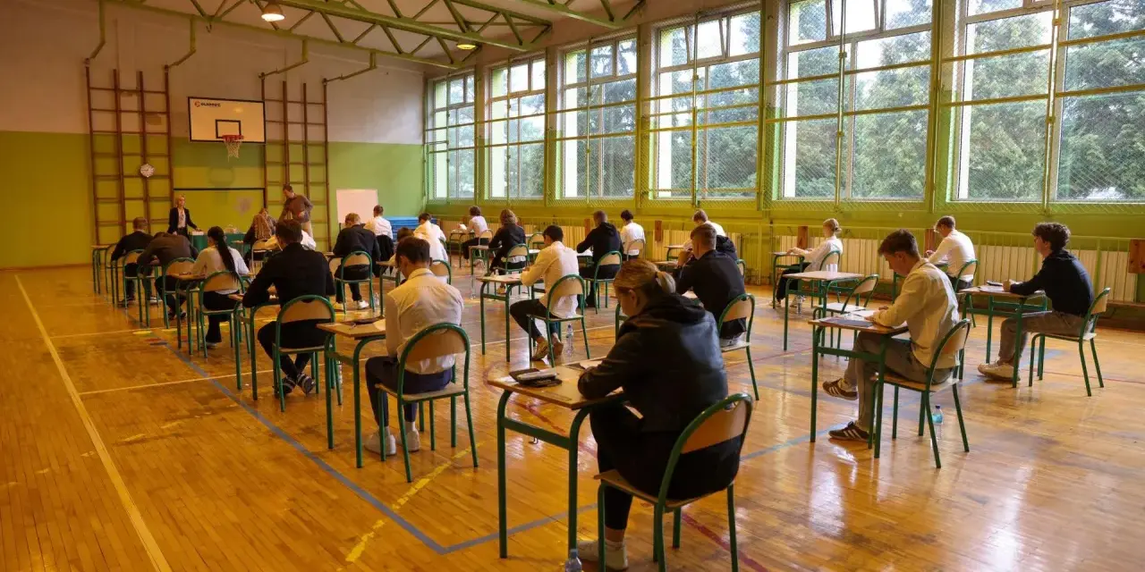 Uczniowie siedzą przy ławkach w sali gimnastycznej, pisząc egzamin.