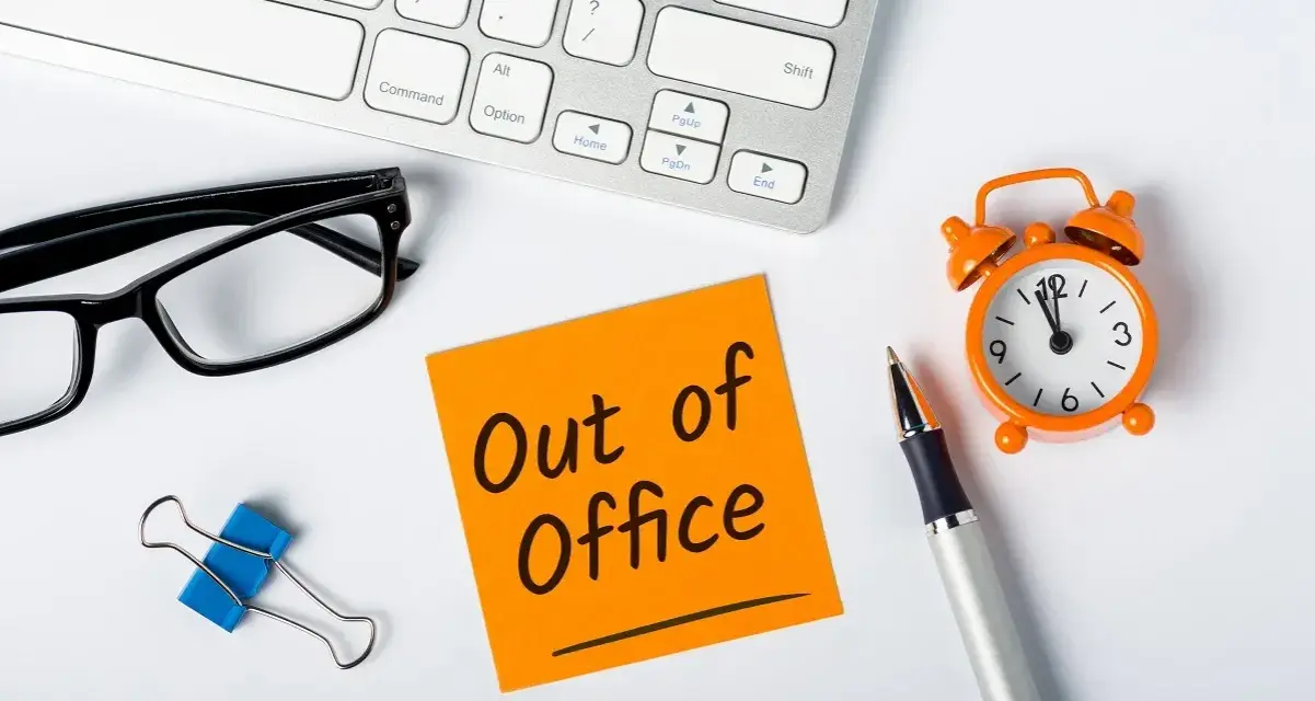 Biurko z klawiaturą, okularami, karteczką "Out of Office", zegarkiem i długopisem.