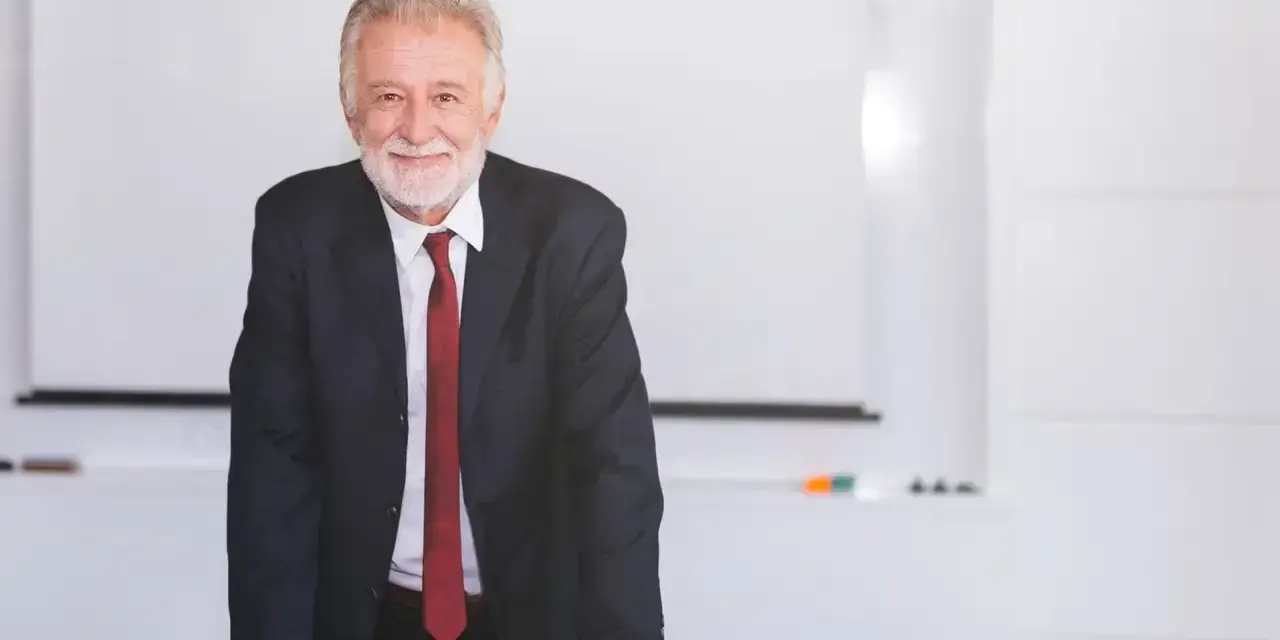 Uśmiechnięty starszy mężczyzna w garniturze stoi przy stole w sali konferencyjnej.