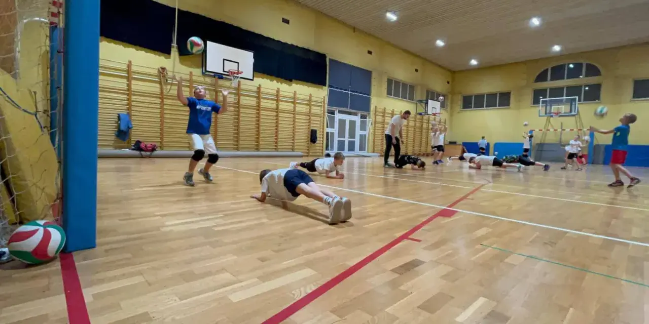 Dzieci ćwiczą siatkówkę na sali gimnastycznej, wykonując pompki i podbijając piłkę.