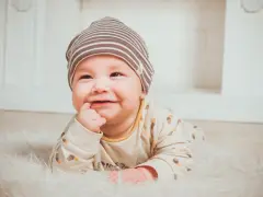 Uśmiechnięte dziecko w czapce leży na białym futerku, wkładając palec do buzi.