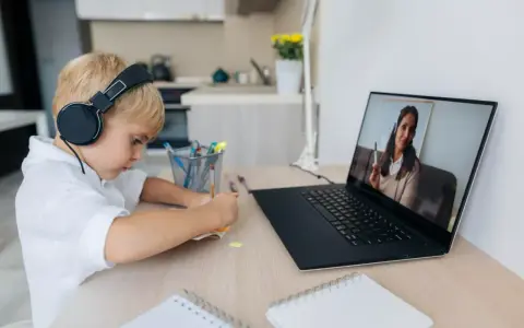 Chłopiec w słuchawkach uczy się zdalnie, patrząc na ekran laptopa z nauczycielem.