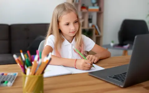 Dziewczynka z blond włosami pisze w zeszycie przy laptopie, obok stoją kolorowe kredki.