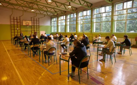 Uczniowie siedzą przy ławkach w sali gimnastycznej, pisząc egzamin.