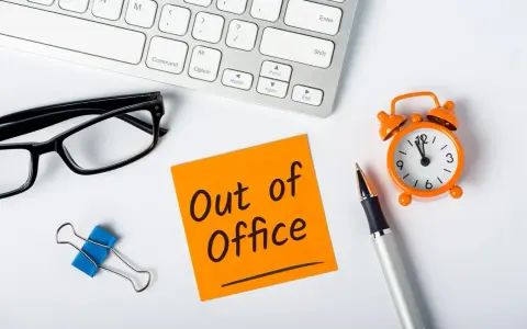 Biurko z klawiaturą, okularami, karteczką "Out of Office", zegarkiem i długopisem.