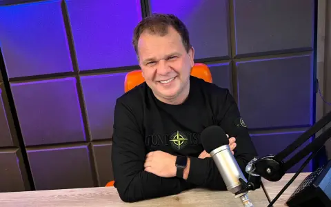 Uśmiechnięty mężczyzna w czarnej bluzie siedzi przy biurku z mikrofonem, na tle fioletowej ściany.