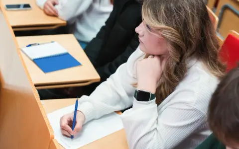 Studentka pisze notatki na wykładzie, opierając podbródek na dłoni.