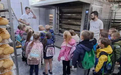 Dzieci z plecakami oglądają piec piekarniczy i półki z chlebem.
