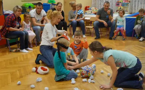 Zajęcia integracyjne z rodzicami w przedszkolu: dzieci z zawiązanymi oczami szukają prezentu, rodzice obserwują.