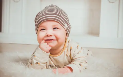 Uśmiechnięte dziecko w czapce leży na białym futerku, wkładając palec do buzi.