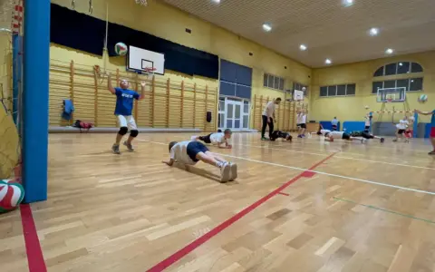Dzieci ćwiczą siatkówkę na sali gimnastycznej, wykonując pompki i podbijając piłkę.