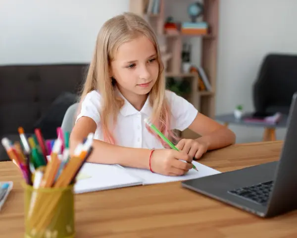 Dziewczynka z blond włosami pisze w zeszycie przy laptopie, obok stoją kolorowe kredki.
