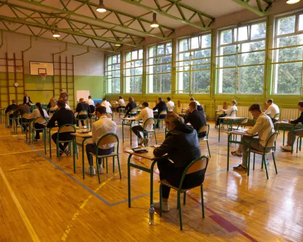 Uczniowie siedzą przy ławkach w sali gimnastycznej, pisząc egzamin.