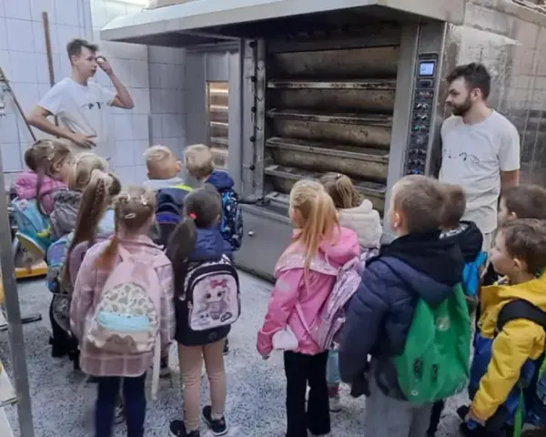Dzieci z plecakami oglądają piec piekarniczy i półki z chlebem.
