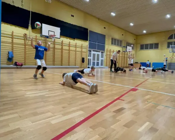 Dzieci ćwiczą siatkówkę na sali gimnastycznej, wykonując pompki i podbijając piłkę.