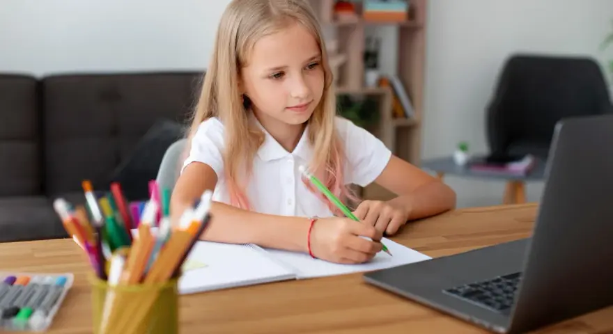 Dziewczynka z blond włosami pisze w zeszycie przy laptopie, obok stoją kolorowe kredki.
