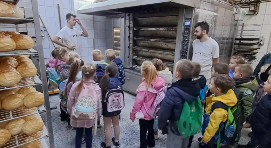 Dzieci z plecakami oglądają piec piekarniczy i półki z chlebem.