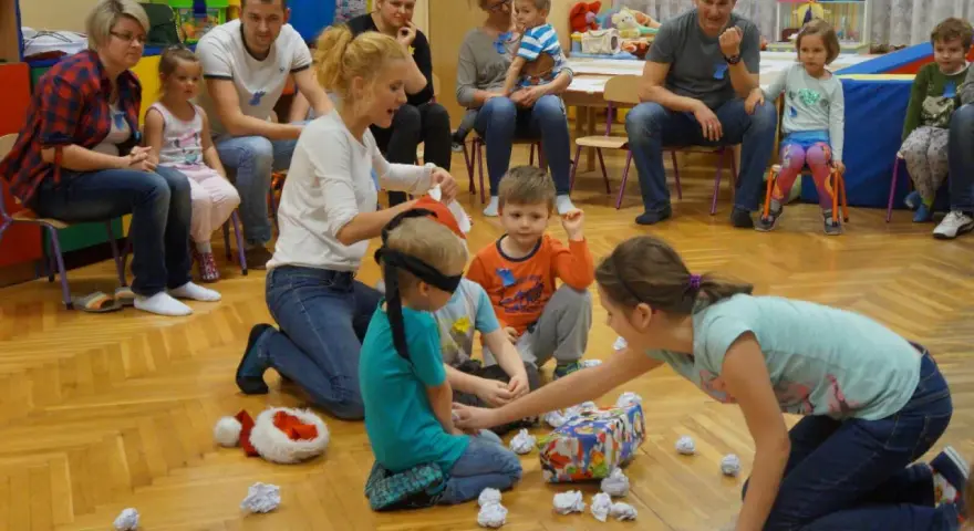 Zajęcia integracyjne z rodzicami w przedszkolu: dzieci z zawiązanymi oczami szukają prezentu, rodzice obserwują.