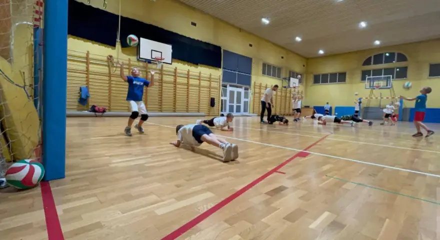 Dzieci ćwiczą siatkówkę na sali gimnastycznej, wykonując pompki i podbijając piłkę.